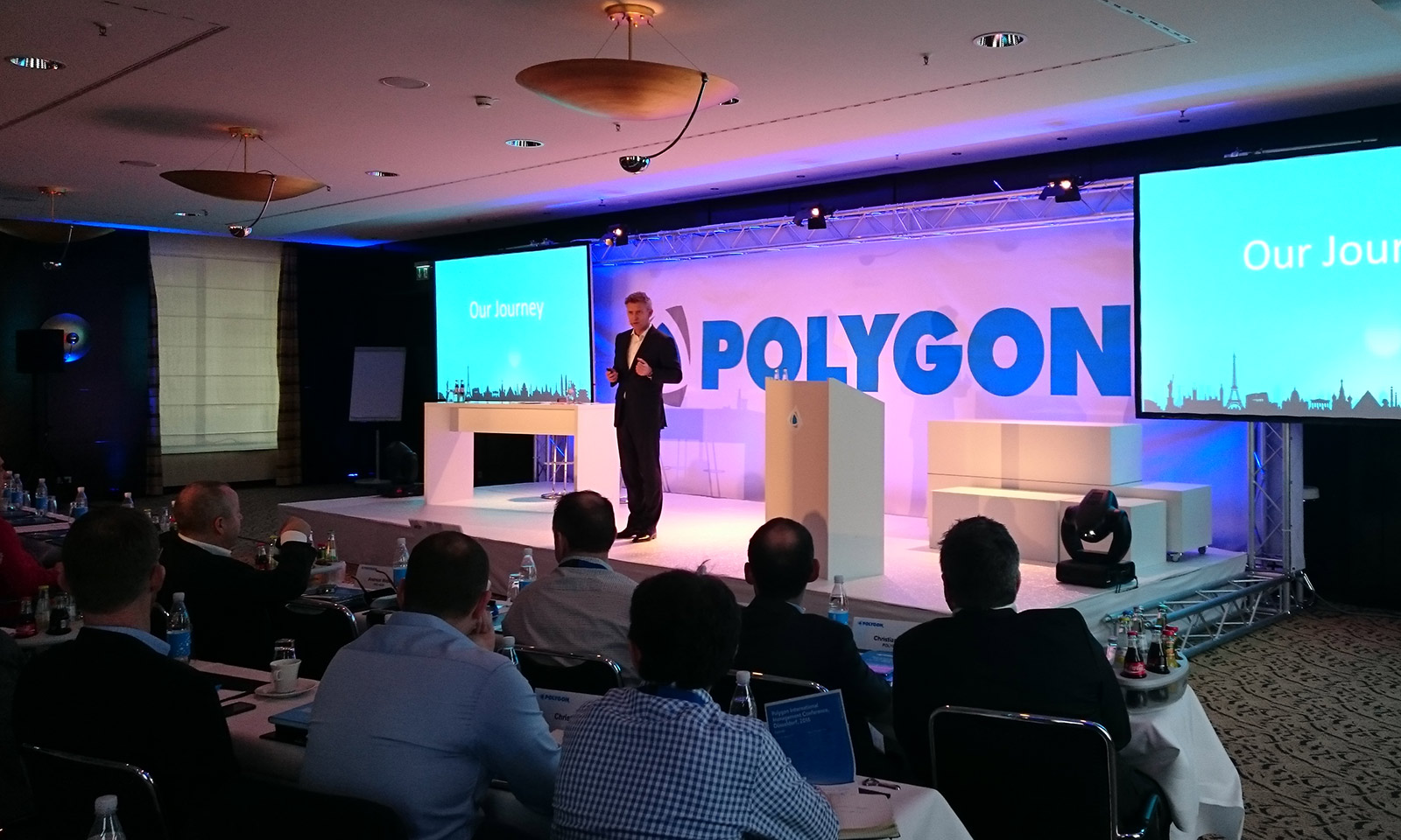 Konferenzsituation mit einem Sprecher auf einer beleuchteten Bühne vor großem POLYGON-Branding, flankiert von zwei Präsentationsscreens und beobachtet von einem sitzenden Publikum