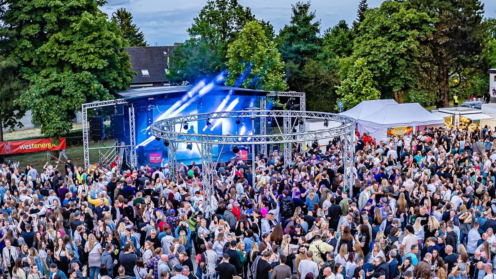 „Großes Open-Air-Festival mit Publikum vor einer beleuchteten Bühne und kreisförmigem Traversensystem, umgeben von Bäumen und Food-Ständen