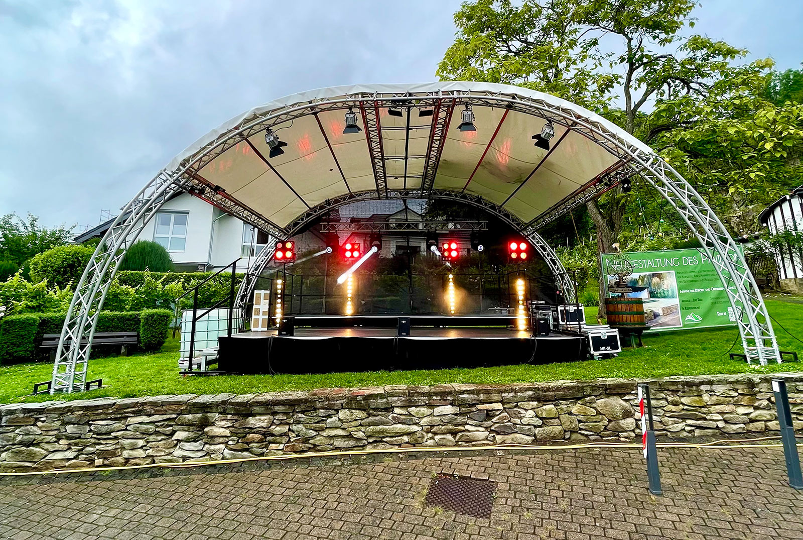 Überdachte Outdoor-Bühne mit halbrundem Traversendach und eingeschalteter Beleuchtung, aufgestellt in einer Gartenanlage vor einer niedrigen Steinmauer