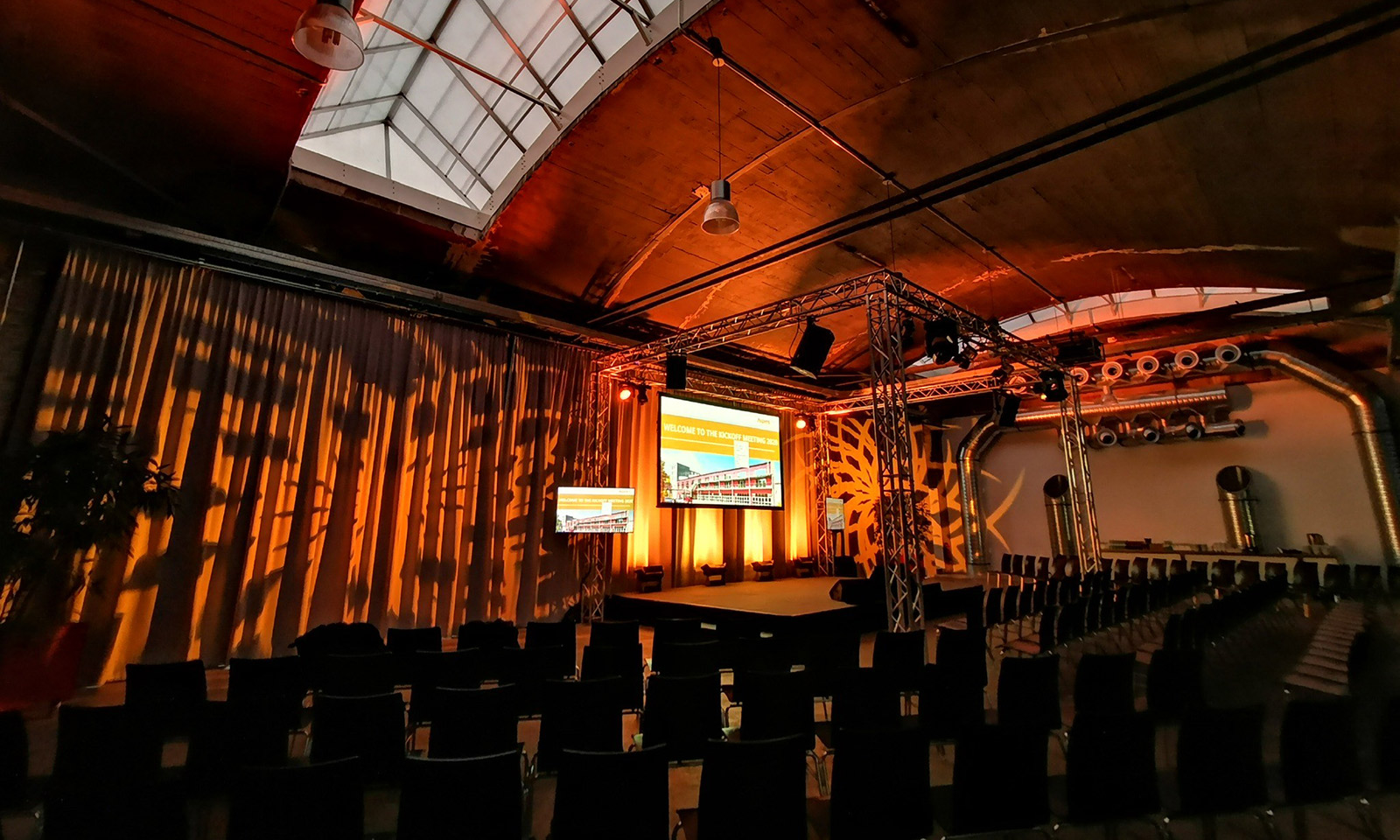 Stimmungsvoll ausgeleuchteter Veranstaltungsraum mit orangefarbener Bühnenbeleuchtung, großem Traversengerüst, Projektionsscreens und bestuhltem Zuschauerbereich