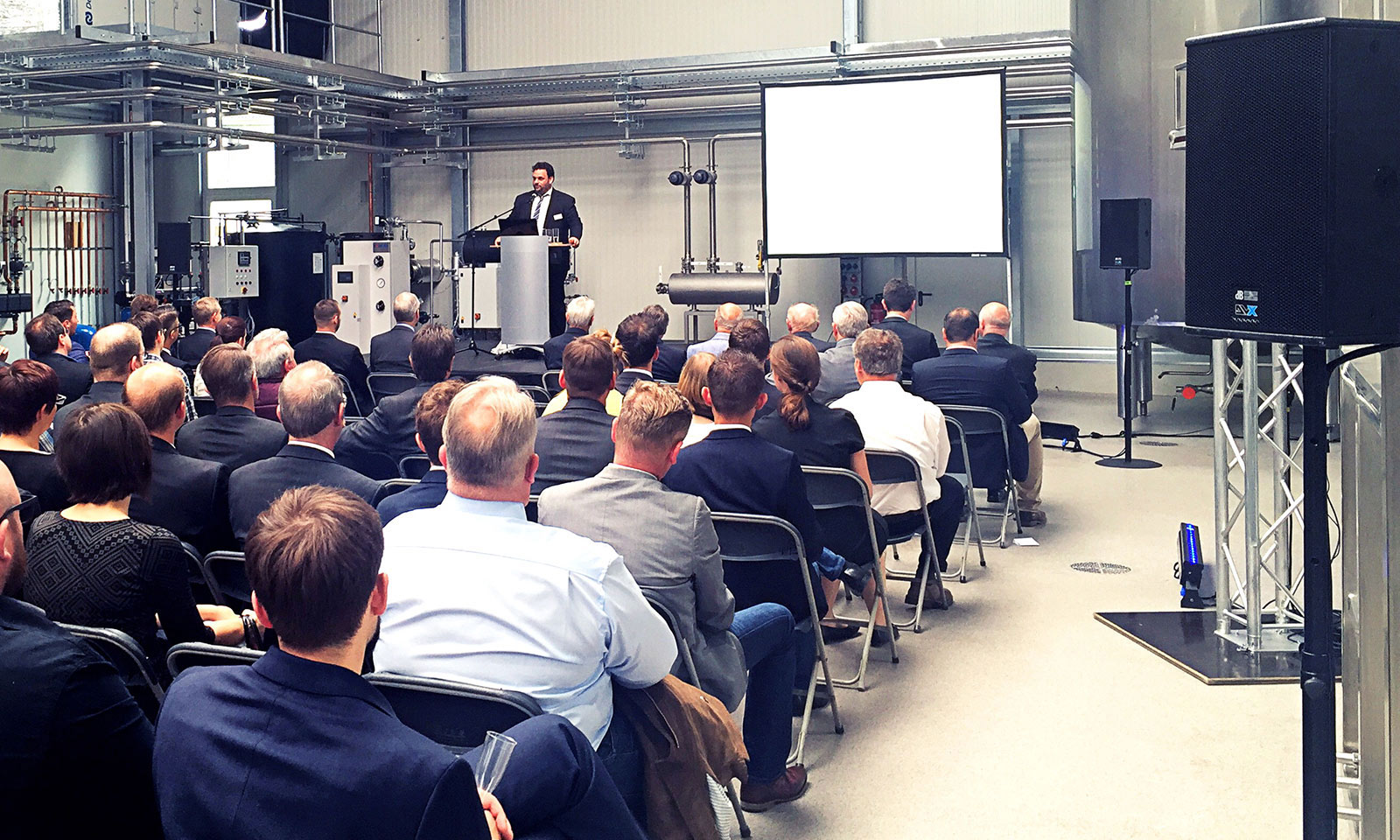 Fachveranstaltung in einer Industriehalle, bei der ein Redner am Podium vor einem sitzenden Publikum präsentiert; im Hintergrund Maschinen und eine Projektionsfläche