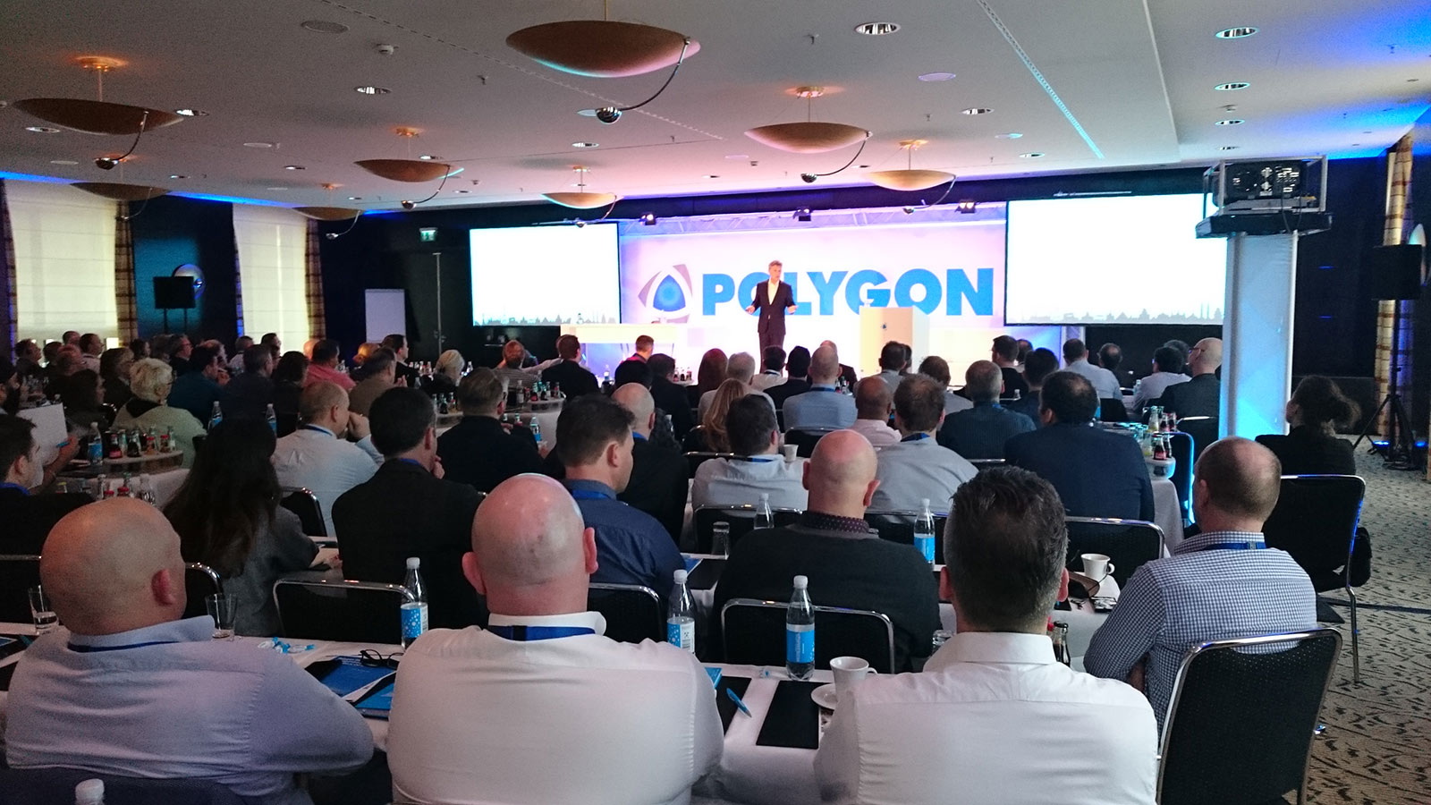 Großer Konferenzraum mit dicht besetzten Tischen, während ein Sprecher auf der Bühne vor einer beleuchteten Polygon-Leinwand präsentiert