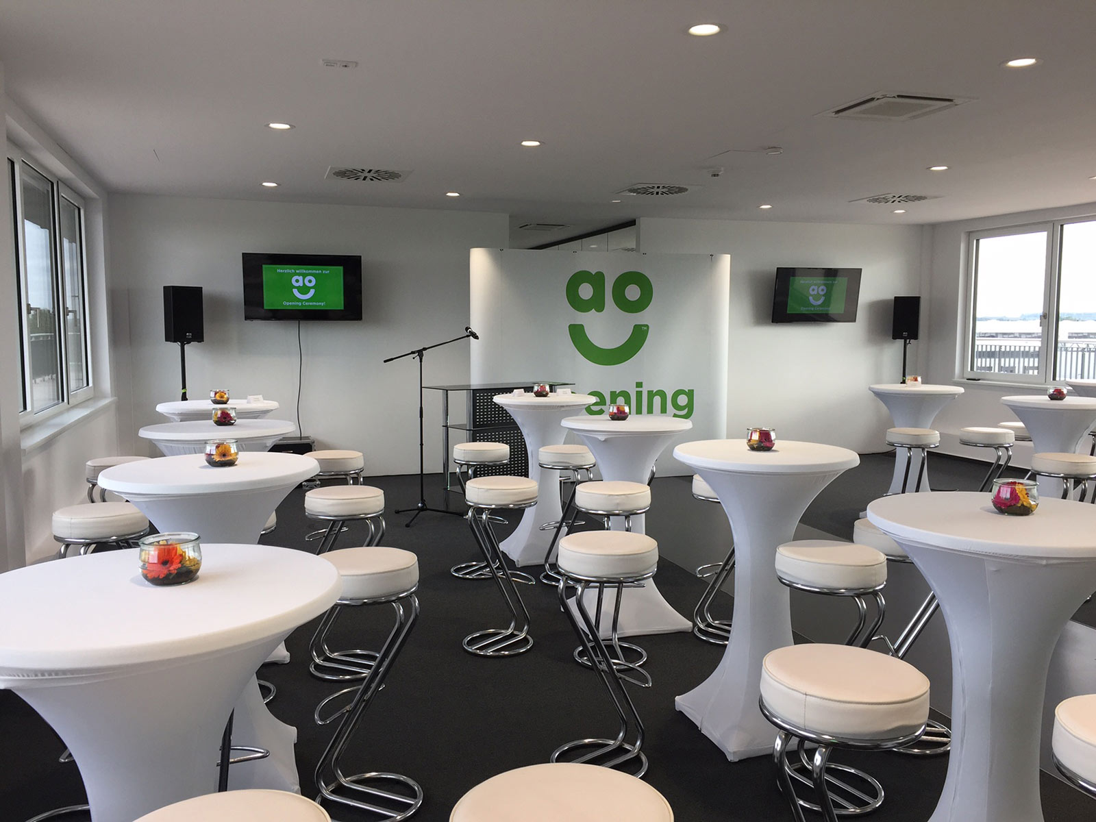 Heller Veranstaltungsraum mit Stehtischen und Barhockern, dekoriert mit kleinen Kerzengläsern, sowie Bühne mit Mikrofon