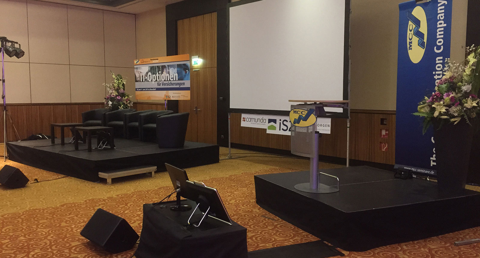 Konferenzbühne mit Podium, Leinwand, Sitzgruppe und Blumenarrangements, vorbereitet für eine Fachveranstaltung
